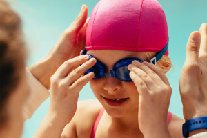 A swim coach helping a little girl put on her swim goggles.